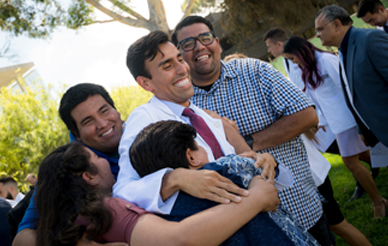 A medical student in a white coat is surrounded by family members who are hugging him