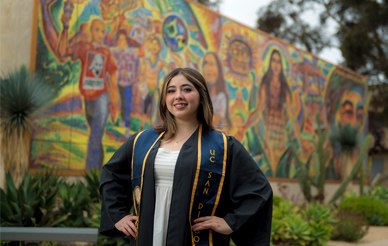Portrait of Letzy Vargas wearing graduation cap and gown in front of a colorful mural