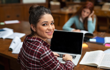 A young woman smiles at the camera while sitting in a classroom with a laptop